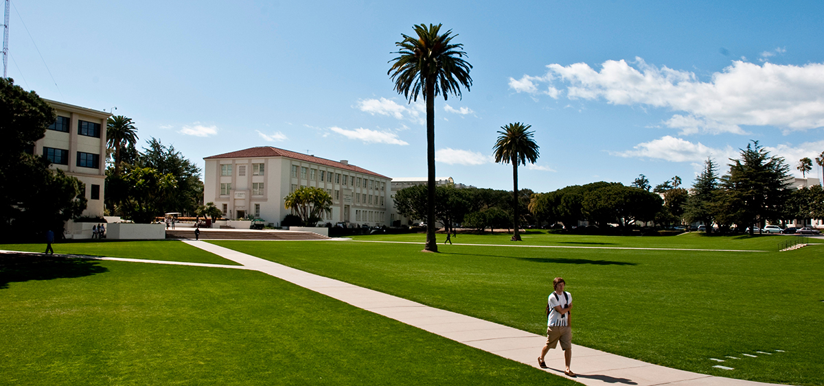 Image of Sunken Garden with St. Robert's Hall in the background. Two palm trees are in the foreground as well as a few students walking through the garden and the surrounding area.